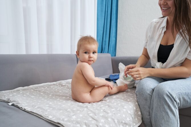 Naked Baby Sitting On Sofa Near Smiling Mother