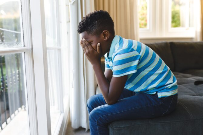 Sad African American Boy Covering His Face And Sitting At Window In Living Room