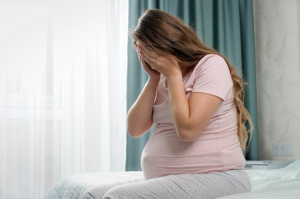 Sad Crying Pregnant Woman Suffering From Depression Sitting On Bed And Holding Her Head. Concept Of